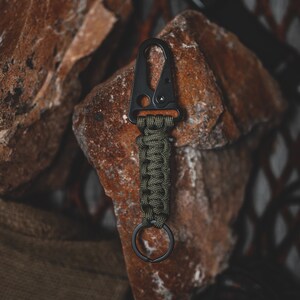 May include: A black metal carabiner with a green paracord wrapped around it. The carabiner has a small ring attached to it.