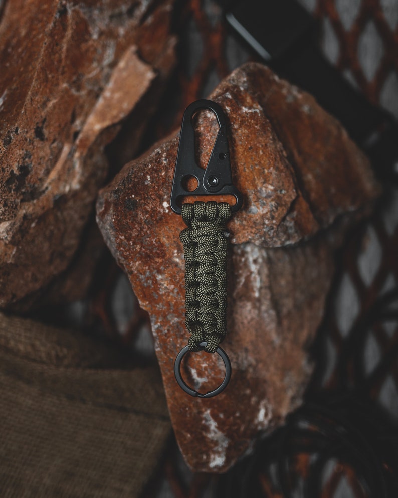 May include: A black metal carabiner with a green paracord wrapped around it. The carabiner has a small ring attached to it.