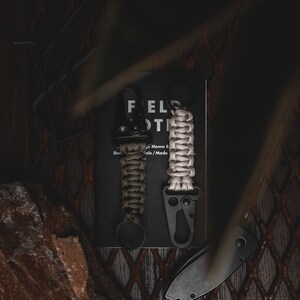 May include: Two black keychains with green and beige paracord wrapped around them. The keychains are attached to a black Field Notes memo book.