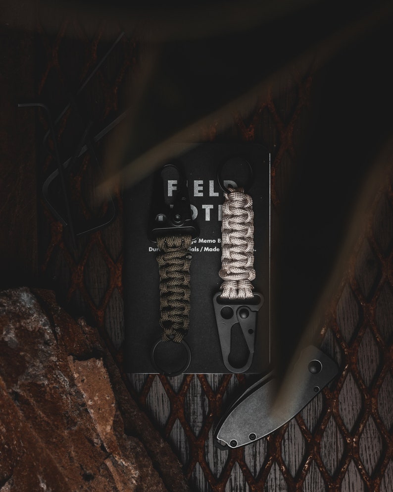 May include: Two black keychains with green and beige paracord wrapped around them. The keychains are attached to a black Field Notes memo book.