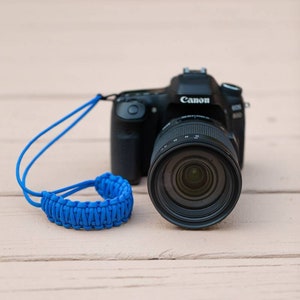 May include: A black Canon camera with a blue braided camera strap. The camera has a large lens and the Canon logo is visible on the top of the camera.