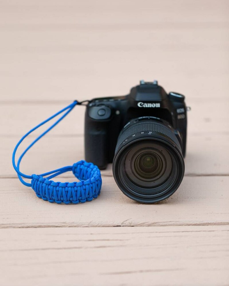 May include: A black Canon camera with a blue braided camera strap. The camera has a large lens and the Canon logo is visible on the top of the camera.