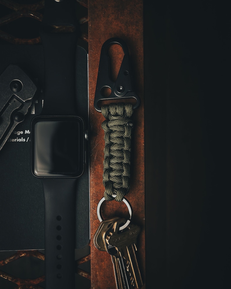 May include: A black metal carabiner with a green paracord wrapped around it. The carabiner is attached to a silver key ring with a set of keys. The carabiner and keys are sitting on a brown leather surface.