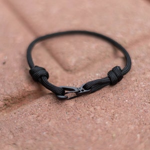 Pode incluir: Um bracelete de paracord preto com um fecho de metal preto. O bracelete &eacute; amarrado com um n&oacute; em cada extremidade.