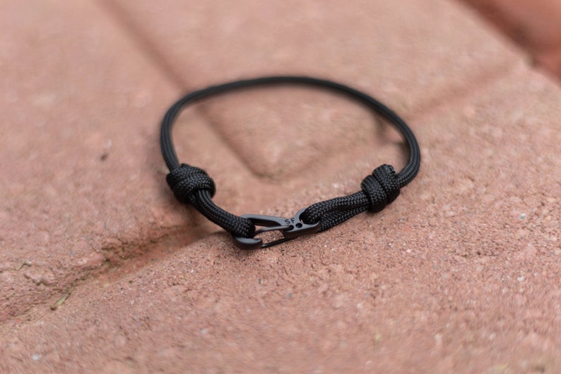 Pode incluir: Um bracelete de paracord preto com um fecho de metal preto. O bracelete &eacute; amarrado com um n&oacute; em cada extremidade.