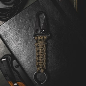 May include: A black metal key chain with a green paracord wrap and a metal ring. The key chain is lying on a black leather surface.