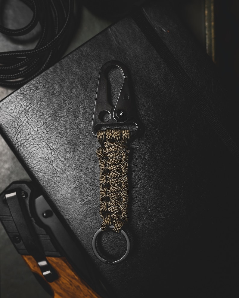 May include: A black metal key chain with a green paracord wrap and a metal ring. The key chain is lying on a black leather surface.