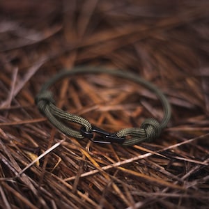 Pode incluir: Um bracelete de paracord verde com um fecho de metal preto. O bracelete est&aacute; sobre um leito de pinhas marrons.
