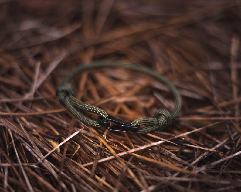 Pode incluir: Um bracelete de paracord verde com um fecho de metal preto. O bracelete est&aacute; sobre um leito de pinhas marrons.