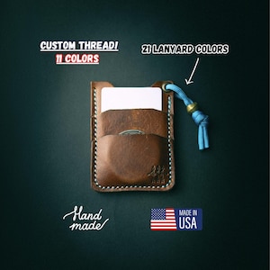 Op de afbeelding: Een bruine leren kaarthouder met een gestikte rand en een blauwe lanyard. De kaarthouder heeft een witte kaart erin en de tekst "Hand made" en "Made in USA" is zichtbaar.
