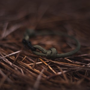Pode incluir: Um bracelete de paracord verde com um fecho preto, deitado sobre um leito de pinhas marrons.