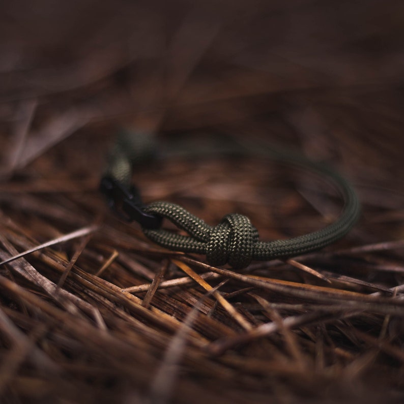 Pode incluir: Um bracelete de paracord verde com um fecho preto, deitado sobre um leito de pinhas marrons.
