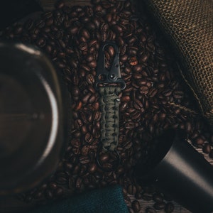 May include: A black and green paracord keychain with a black metal carabiner clip. The keychain is lying on a bed of coffee beans.