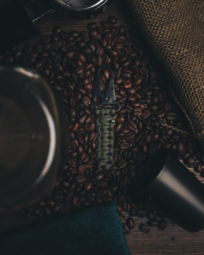 May include: A black and green paracord keychain with a black metal carabiner clip. The keychain is lying on a bed of coffee beans.