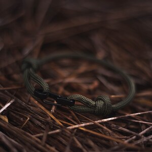 Pode incluir: Um bracelete de paracord verde com um fecho de metal preto. O bracelete est&aacute; sobre um leito de pinhas marrons.