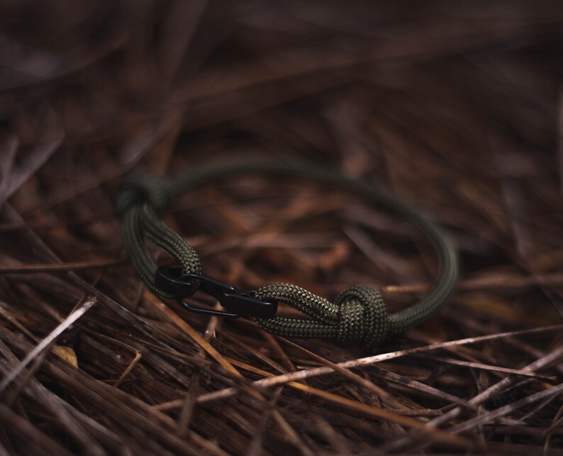 Pode incluir: Um bracelete de paracord verde com um fecho de metal preto. O bracelete est&aacute; sobre um leito de pinhas marrons.