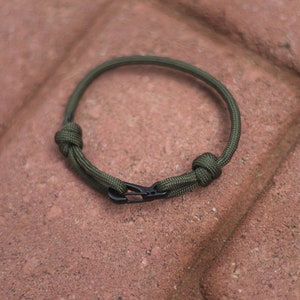Pode incluir: Um bracelete de paracord verde com um fecho de metal preto. O bracelete &eacute; amarrado com um n&oacute; em cada extremidade.