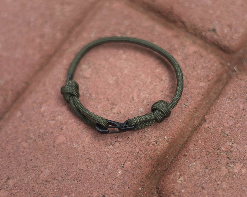 Pode incluir: Um bracelete de paracord verde com um fecho de metal preto. O bracelete &eacute; amarrado com um n&oacute; em cada extremidade.