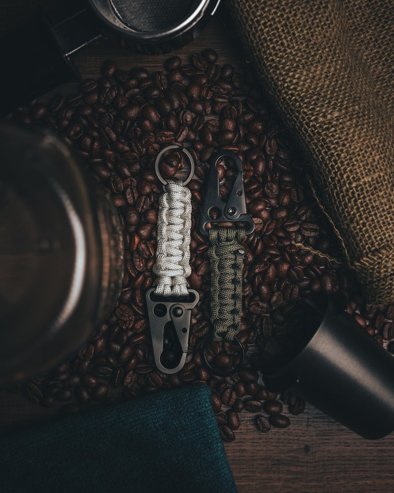 May include: Two black paracord keychains with metal clips, one white and one green, on a bed of coffee beans.