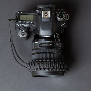 May include: A black Canon EOS camera with a 24-105mm lens attached. The camera has a black strap and a black lens cap. The camera is on a gray surface.