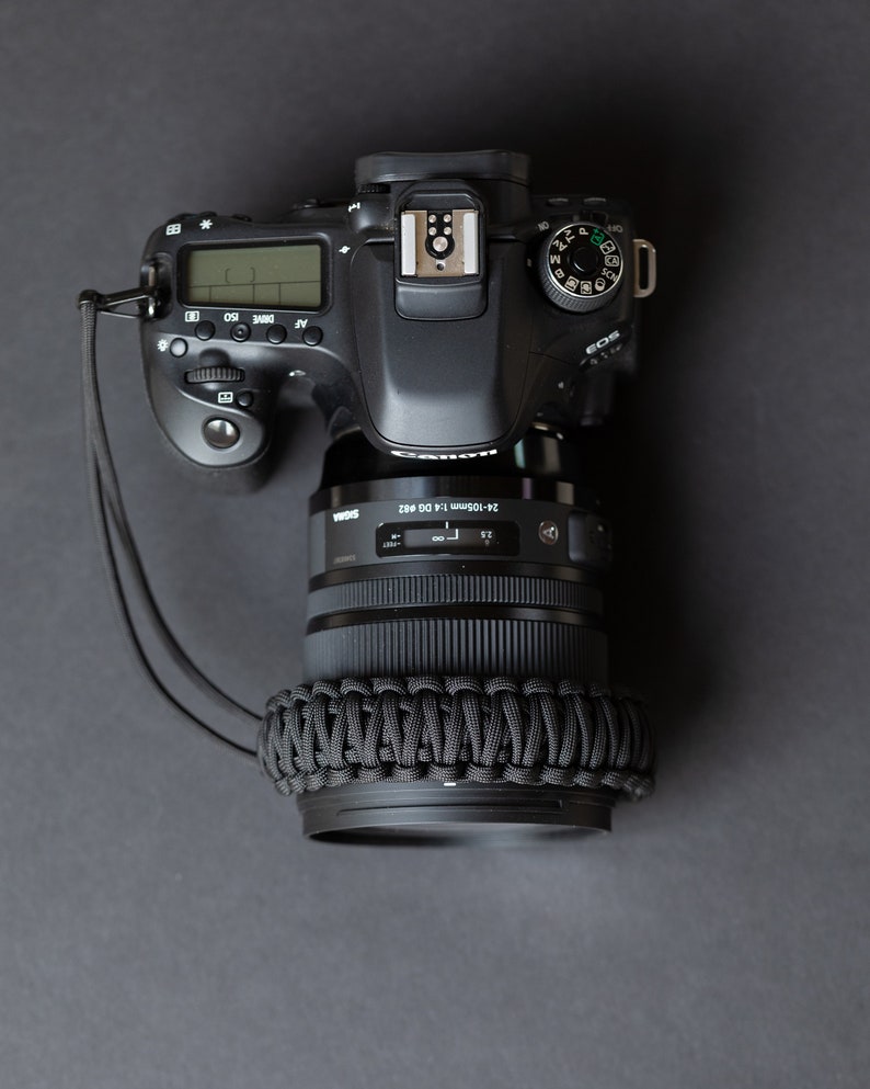 May include: A black Canon EOS camera with a 24-105mm lens attached. The camera has a black strap and a black lens cap. The camera is on a gray surface.