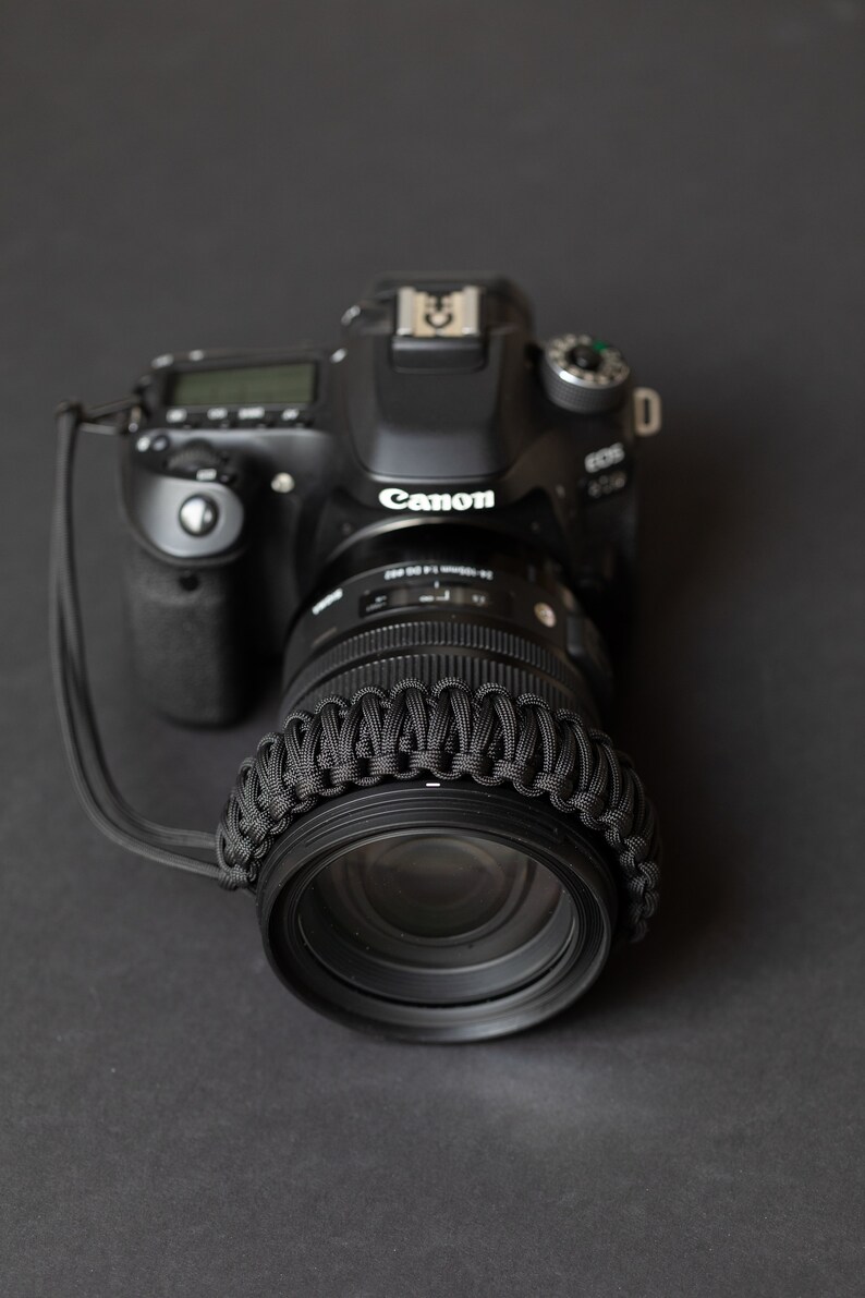 May include: A black Canon DSLR camera with a black lens. The lens has a black paracord strap around it.