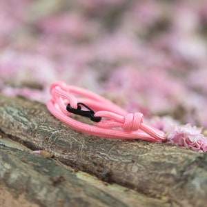 Könnte beinhalten: Ein pinkfarbenes Paracord-Armband mit einem schwarzen Plastikverschluss. Das Armband liegt auf einem Stück verwittertem Holz.