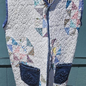May include: Quilted vest with a patchwork design featuring various colors and patterns. The vest has a blue trim, two denim pockets with decorative stitching, and a pink hem. It is displayed on a metal form.