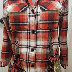 May include: A button-down plaid shirt in red, white, and brown. The shirt features two chest pockets and a patchwork ruffle hem with floral and geometric patterns. The garment is displayed on a mannequin.