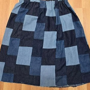May include: A knee-length skirt constructed from a patchwork of rectangular denim fabric pieces in alternating shades of dark indigo and light blue. The garment features a gathered elastic waistband and a straight hemline with visible stitching.