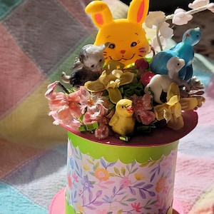 May include: A pink and green hatbox with a floral pattern. The hatbox is filled with a variety of Easter decorations, including a yellow bunny, a blue bird, a white lamb, a yellow chick, and pink and yellow flowers.