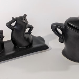 May include: A set of three black ceramic teapots with whimsical designs. Two of the teapots are on a matching black ceramic tray. The teapots have lids and handles.