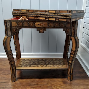 May include: A wooden game table with intricate inlaid designs. The table has a hinged top that opens to reveal a playing surface. The table has four legs with a curved design.