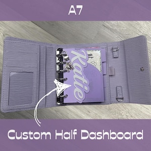 May include: A light purple A7 binder with a textured finish, featuring a metal ring mechanism and a custom half dashboard insert. The dashboard is light purple with the name Katie in white script and a small silver butterfly charm attached.