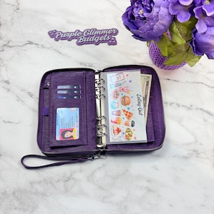 May include: A purple zippered binder wallet with a wrist strap, featuring interior card slots, a clear ID window, and a six-ring metal mechanism. It contains a transparent divider printed with food illustrations and the text Eating Out.