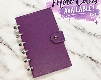 May include: A purple disc-bound planner with a textured cover and a snap button closure. The side features silver-colored metal discs for binding. The text "More Colors AVAILABLE!" is printed in purple script above the planner.