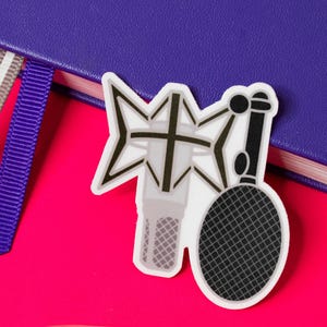 Puede incluir: Una pegatina con un diseño de micrófono y un filtro antipop en forma de estrella. Está colocada sobre una superficie rosa brillante junto a un cuaderno morado con dos cintas de marcador. El micrófono es gris y negro.