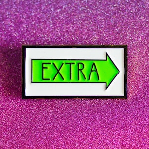 May include: A white enamel pin with a black border and a green arrow pointing to the right. The arrow has the word "EXTRA" written in black inside of it.