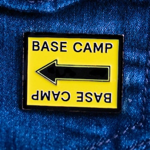 Könnte beinhalten: Rechteckiger Emaille-Pin mit gelbem Hintergrund und schwarzem Rand. Der Pin zeigt die Worte "BASE CAMP" über einem schwarzen Pfeil, der nach links zeigt, und die Worte "BASE CAMP" darunter.
