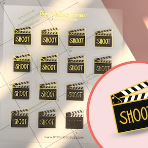 May include: A sheet of clear stickers with gold foil accents. Each sticker features a black and white clapperboard with the word "SHOOT" in gold lettering. The stickers are arranged in a grid pattern on a white background.