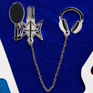 Könnte beinhalten: Silberfarbene Brosche mit Tennisschläger, Mikrofon und Kopfhörern, verbunden durch eine Kette. Die Brosche hat schwarze Akzente und ist vor blauem Hintergrund platziert. Das Design deutet auf ein Thema aus Musik und Sport hin.