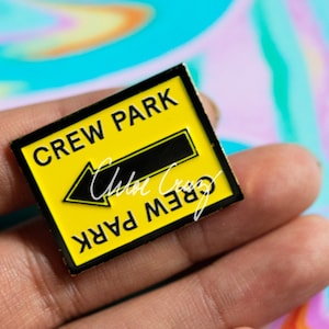 May include: A yellow and black enamel pin with a black arrow pointing left and the text "CREW PARK" in black letters.