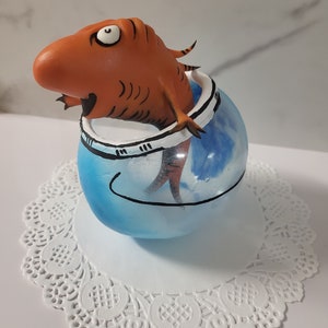 May include: A whimsical orange fish character emerging from a blue and white egg-shaped container. The container is decorated with a black outline and rests on a white doily. The fish has a surprised expression.