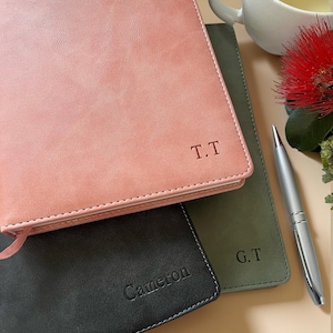 May include: Three personalised notebooks in pink, green, and black. The pink notebook has the initials "T.T." embossed, the green notebook has "G.T.", and the black notebook has "Cameron." A silver pen rests beside the notebooks, with a cup and flower in the background.