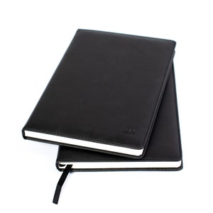 May include: Two black leather-bound journals with white edges and a black ribbon bookmark. The top journal has the initials "SN" embossed on the front cover.