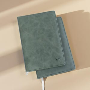 Puede incluir: Se muestran dos cuadernos verde salvia con una cubierta texturizada. El cuaderno superior tiene las iniciales "N.S" impresas en la esquina inferior derecha. Cada cuaderno tiene una cinta blanca como marcador. Los cuadernos están sobre una superficie de color beige claro.