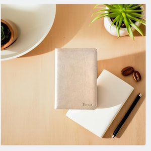 May include: A gold-coloured leather notebook with the name "Bonnie" embossed on the front cover. The notebook is lying on a wooden surface next to a white notebook and a black pen.