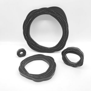 Puede incluir: Colección de cinco objetos negros abstractos en forma de anillo. Los anillos varían en tamaño, con un marco circular grande y formas más pequeñas e irregulares. El diseño presenta un patrón tejido en capas, creando una apariencia texturizada. Sobre un fondo blanco.