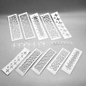 May include: A selection of white paper cut-out designs, including rectangular bookmarks and decorative border strips. The designs feature various patterns, such as circles, floral motifs, and cross shapes, creating a delicate and intricate aesthetic.