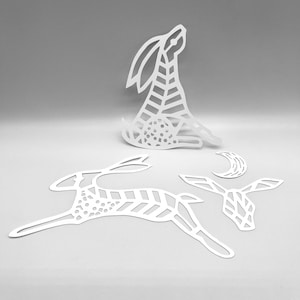 Pode incluir: Recortes de papel branco de coelhos e uma lua crescente. Os coelhos estão em várias poses, incluindo sentados e saltando. Os desenhos apresentam padrões geométricos e espaços abertos, criando uma estética delicada e arejada.
